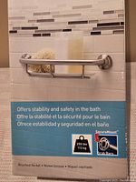 Photo showing the grab bar mounted on tiled wall with soap and sponge, displaying its brushed nickel finish and integrated shelf, labeled with product safety and weight capacity.