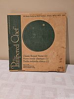 Photo of the unopened cardboard box with Pampered Chef branding and a circular cutout showing the pizza stone inside.