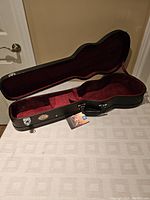 Open black hard case with plush red interior shaped to fit a baritone ukulele, showing interior storage compartment and Profile badge inside.