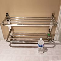 Front view of the stainless steel towel rack showing two shelves made of horizontal bars and supporting vertical brackets mounted on a table against a beige wall with a water bottle for size reference.