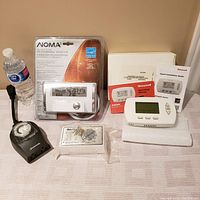 Lot items arranged on table with water bottle for scale: indoor/outdoor timer, sealed Noma programmable thermostat, Honeywell thermostat with manuals, new humidifier control.