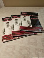 Three packaged men's undershirts displayed on a table, showing the front of package with a print image of a male model wearing the undershirt and branding.