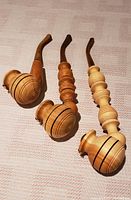 Three cherrywood pipes of varying sizes arranged side by side on a beige fabric surface, showing their curved stems and rounded bowls with black lines.
