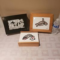 Three wooden and ceramic boxes arranged side by side. Left black wooden box with silver-tone raised Haida design signed by Clarence A. Wells. Center wooden framed ceramic tile with orca design. Front wooden box with sliding ceramic lid featuring salmon design.
