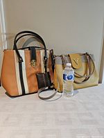 Two handbags placed side by side with a water bottle for scale on table. Caramel color bag has stripes and a gold clasp, yellow bag has large 'V' logo on front flap.