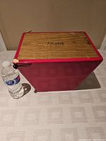 Bright red Tycoon Percussion cajon drum with wood grain playing surface and logo on top, water bottle for scale against beige background, showing minor wear and paint chipping on edges.