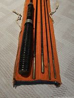 Folded four segments of vintage fly fishing rod with black ribbed handle, brass and steel ferrules, inside orange fabric carrying case with white drawstrings.