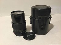 Canon 135mm lens with leather case and Vivitar lens cap, showing frontal view of lens with markings and case beside it.