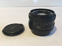 Front and side view of Canon FD 50mm f/1.8 lens with front lens cap off and placed beside the lens, showing aperture ring and distance markings.