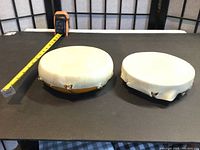 Two small handcrafted First Nation style drums made from wood bowl bases with white cured hide stretched over. Photo shows the drums from a perspective angle with tape measure for size reference.