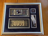 Framed collection with three main items mounted on dark blue matting inside gold and white frame, includes two tickets, a pin, and an event tag plaque.