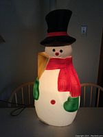 Front view of illuminated Frosty the Snowman blow mold sitting on chair, glowing light inside, showing black top hat with red band, red scarf, green mittens, yellow broom.