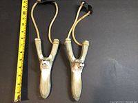 Pair of hand carved wolf face slingshots next to a measuring tape showing length about 7 inches.