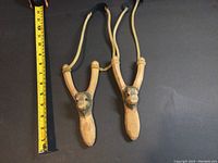 Photo showing two hand carved wooden slingshots with buffalo face designs next to measuring tape indicating length around 8.5 inches.