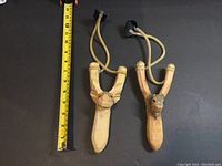 Pair of hand carved wooden slingshots next to measuring tape showing approximate length of 7 inches each.