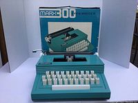 Marx 300 toy typewriter with original box shown behind it. Teal blue body and white keys visible, box shows illustration and branding.
