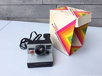 Front view of Polaroid Pronto B camera with shutter button, lens, and viewfinder visible alongside original box.