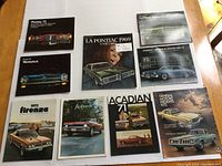 Lot of nine Pontiac automobile sales brochures from late 1960s and early 1970s, laid out on a wooden table. Brochures show multiple Pontiac vehicle models with some covers in French.