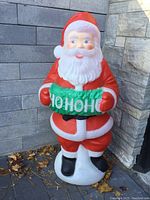 Front view showing Santa holding a green HO HO HO sign, against a gray stone wall with some leaves on the ground.