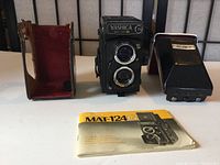 Front view of Yashica Mat-124G flanked by red leather case and prism finder with manual in front