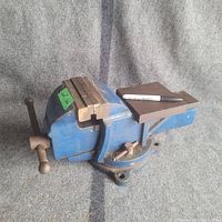 Side angle showing blue painted heavy duty vise with attached anvil and swivel base, resting on gray fabric.