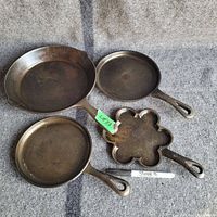 Top view of all four cast iron pans laid flat on grey carpet. Includes three round pans of varying size and one flower-shaped pan. Sharpie marker for scale.