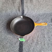 Top-down view of the 9-inch cast iron pan with orange handle on grey fabric background with lot number label.