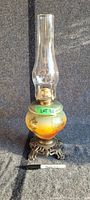 Full view of oil lamp showing glass chimney, painted glass body, and brass ornate base with marker for scale.
