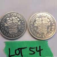 Pair of British Columbia commemorative silver-colored dollar coins side by side with green tag marked 'LOT 54'.