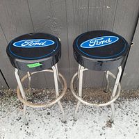 Pair of Ford shop stools side by side showing black padded seats with large Ford logos and chrome metal legs with rust and paint wear, standing on snow-dusted ground against a wall.
