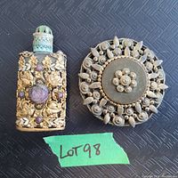 Top view of vintage decorative scent bottle with floral embossing and gem-like stones next to large floral filigree round brooch on black textured background labeled Lot 98