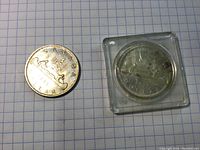 Two 1965 Canadian silver dollar coins, one loose and one in a plastic protective case, placed on grid paper.