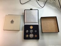 Open presentation case displaying all seven coins in velvet insert with outer cardboard slipcover to left and removed case lid to right