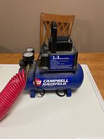 Front view of Campbell Hausfeld air compressor on table with pressure gauges and red coiled hose, showing brand label and motor mounted on blue tank.
