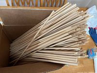 Open box filled with numerous light-colored wooden dowels, each about 2 feet long, stacked inside a brown cardboard box on a table.