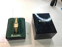 Full view of gold plated Bulova watch in green velvet box next to black glossy outer box.