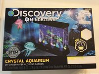 Front of box showing Discovery Mindblown Crystal Aquarium DIY Underwater Glowing Garden kit, age 8+, STEM logo, and crystal aquarium image with glowing hanging crystals and jellyfish graphics.
