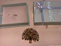 Gold-tone metal peacock brooch with multi-colored rhinestones in a light blue gift box with ribbon.
