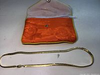 Gold necklace laid out flat showing full length and accompanying orange satin pouch with brand text inside the flap.