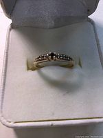 Front-facing view of gold ring with dark central gemstone and diamond accents on white cushion inside ring box.