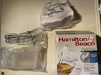 Hamilton Beach hand mixer with beaters, whisk attachments, bowl rest in sealed plastic with box showing front and info.