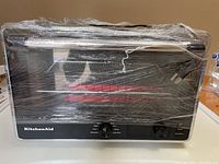 Front view of KitchenAid toaster oven wrapped in plastic showing brand logo and control knobs.