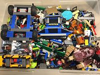 Clear storage bin containing assorted loose Lego pieces: vehicle chassis, wheels, plates, bricks and specialized elements