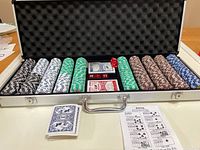 Full poker chip set inside an aluminum lockable case lined with foam. Visible chips in black, white, green, brown, and blue, two decks of playing cards in plastic wrap, and red dice.