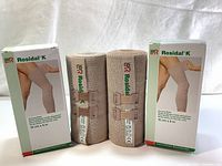 Two Rosidal K bandage rolls with packaging standing upright on fabric background. Beige bandages with Velcro outside packaging boxes with image and product info.