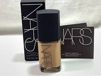 NARS Sheer Glow Liquid Foundation bottle with black matte pump cap and clear glass bottle, front showing brand and product name. Box and inserts placed behind the bottle.