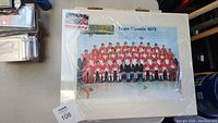 Photo of a 1972 Team Canada hockey team picture with players in red and white jerseys seated and standing in rows, covered by plexiglass with plastic sheet still on it.