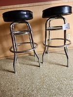 Two chrome and black vinyl leather cushioned bar stools standing upright on carpeted floor against wood panel wall.