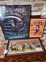 Three framed art prints leaning against a brick wall on a patterned carpet, showing Mike Tyson feeding pigeons, Lenny Bruce poster, and H.R. Giger Alien print.