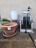 Set of 5 pots (terracotta, ceramic, and plastic) including green ceramic pot with rosemary plant, white plastic pot and terracotta pot. Also shows white hosemobile reel with a garden hose coiled.